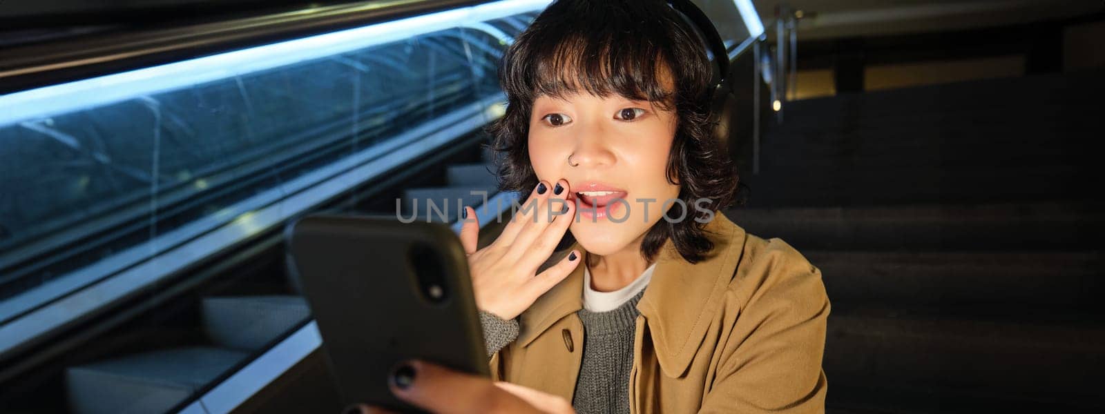 Close up portrait of korean girl looks surprised at mobile phone, reads something amazing on smarpthone screen, watching video in headphones, sits on staircase by Benzoix