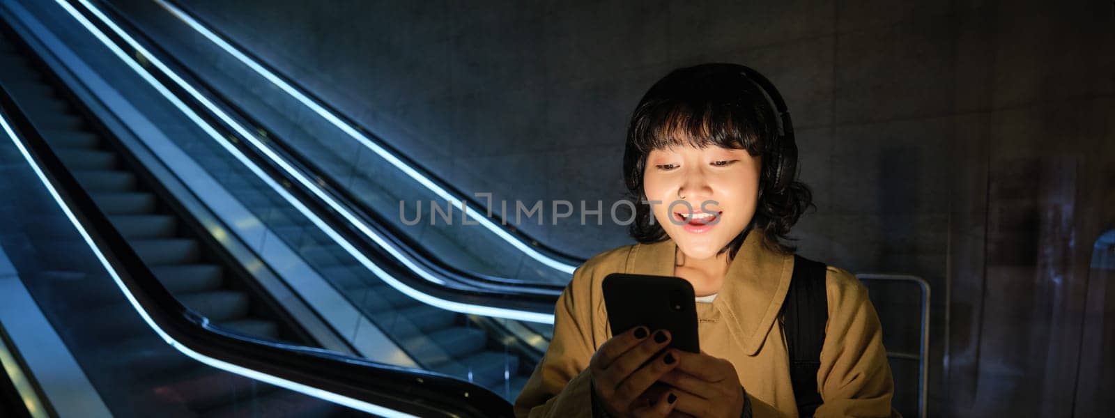Joyful asian woman student, looking surprised and happy at smartphone screen, reading amazing news, listening music in headphones, found smth online on mobile phone app.