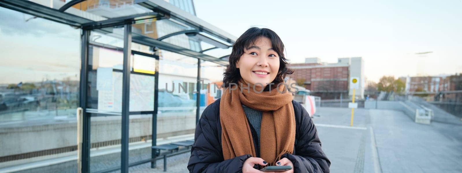 Happy smiling korean girl, using mobile phone, standing on bus stop with smartphone, looking at departure schedule on application, posing in winter clothes by Benzoix