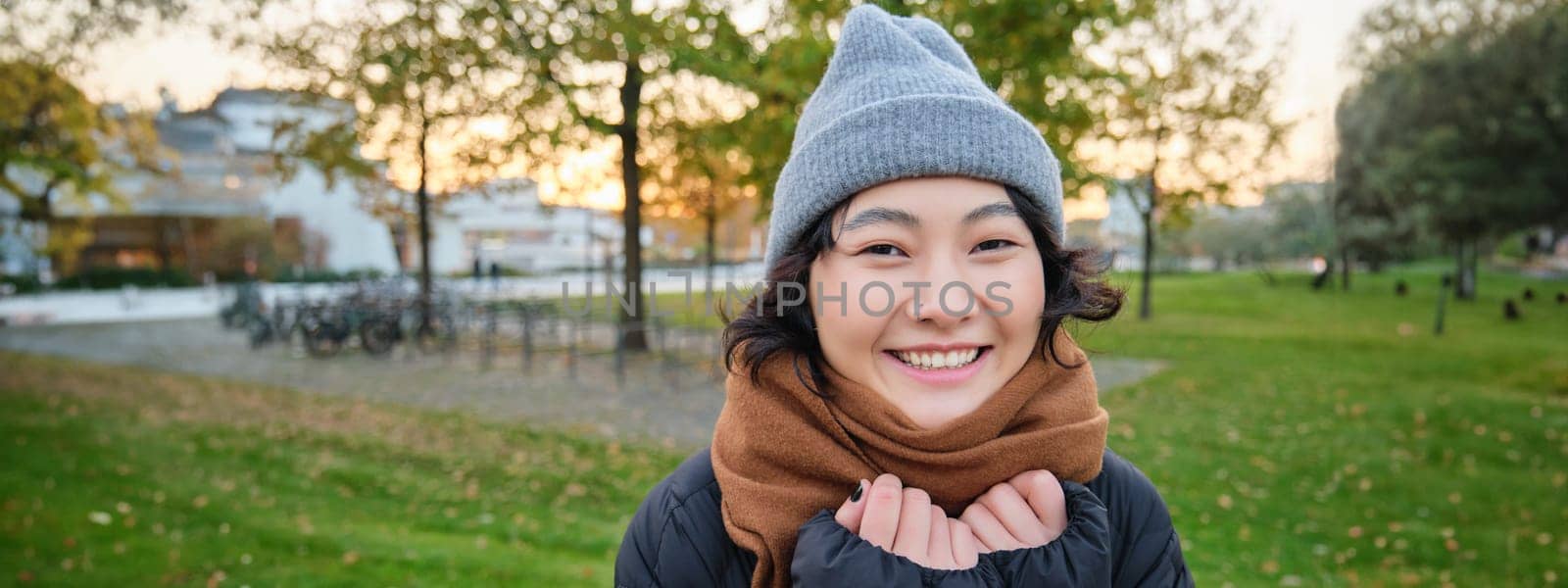 Beautiful young asian woman in warm hat and scarf, smiles at camera, walks around park on chilly spring day, posing with happy face expression.