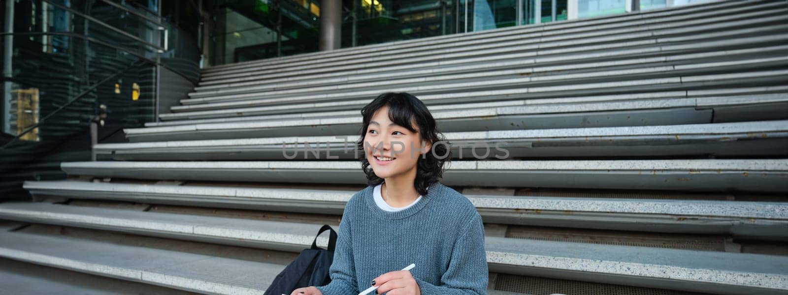 Smiling girl, graphic designer, using digital tablet and pen tool to draw, does home assignment for university, sits on stairs by Benzoix
