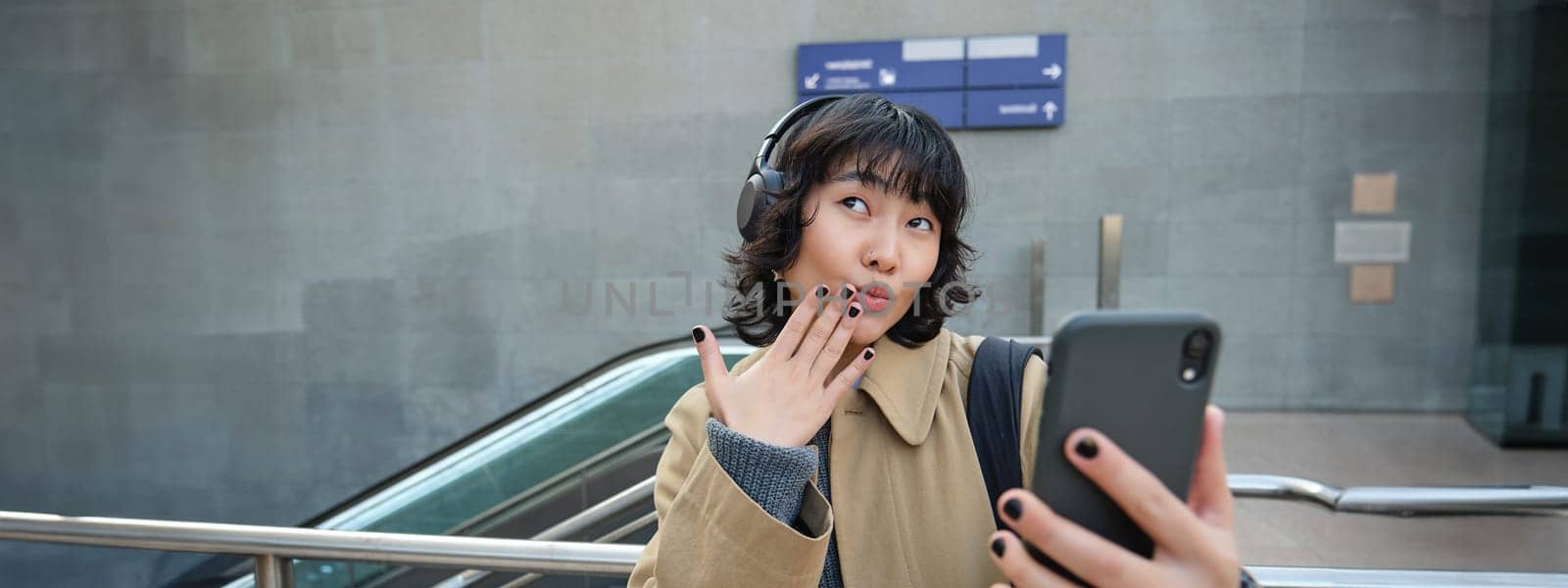 Cute and funny korean girl in headphones, posing for selfie, takes photo on smartphone and looks silly, stands on street in city.