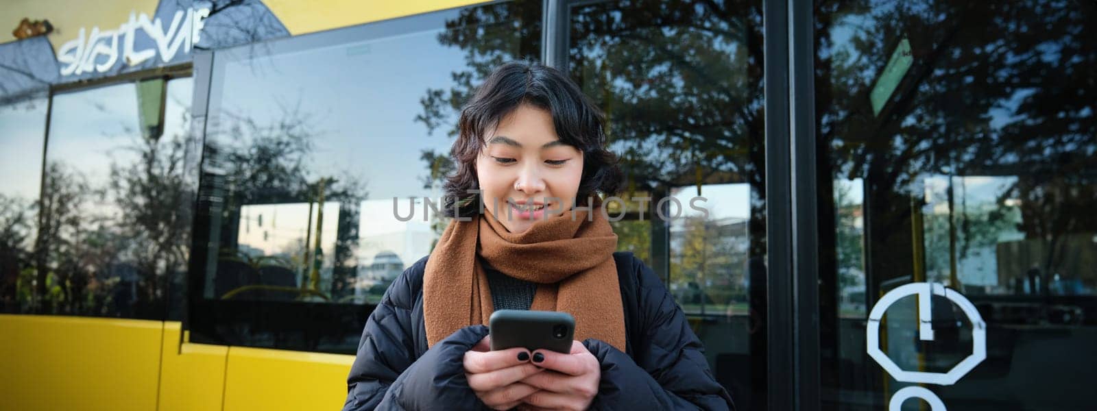 Beautiful korean girl, student on bus stop, looking at her smartphone, checking timetable, reading text message, wearing winter clothes by Benzoix