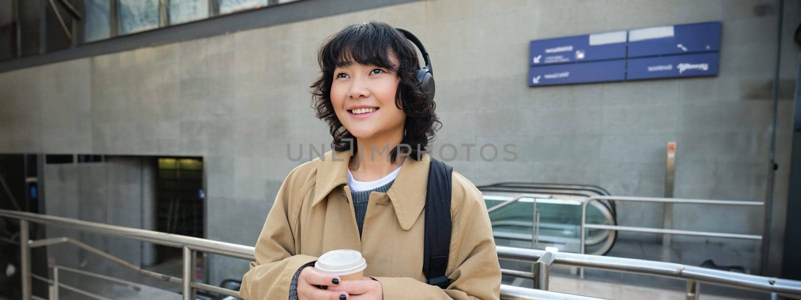 Beautiful smiling Korean girl, drinks takeaway coffee, listens music in headphones, wears trench and backpack, stands on train station, travelling.