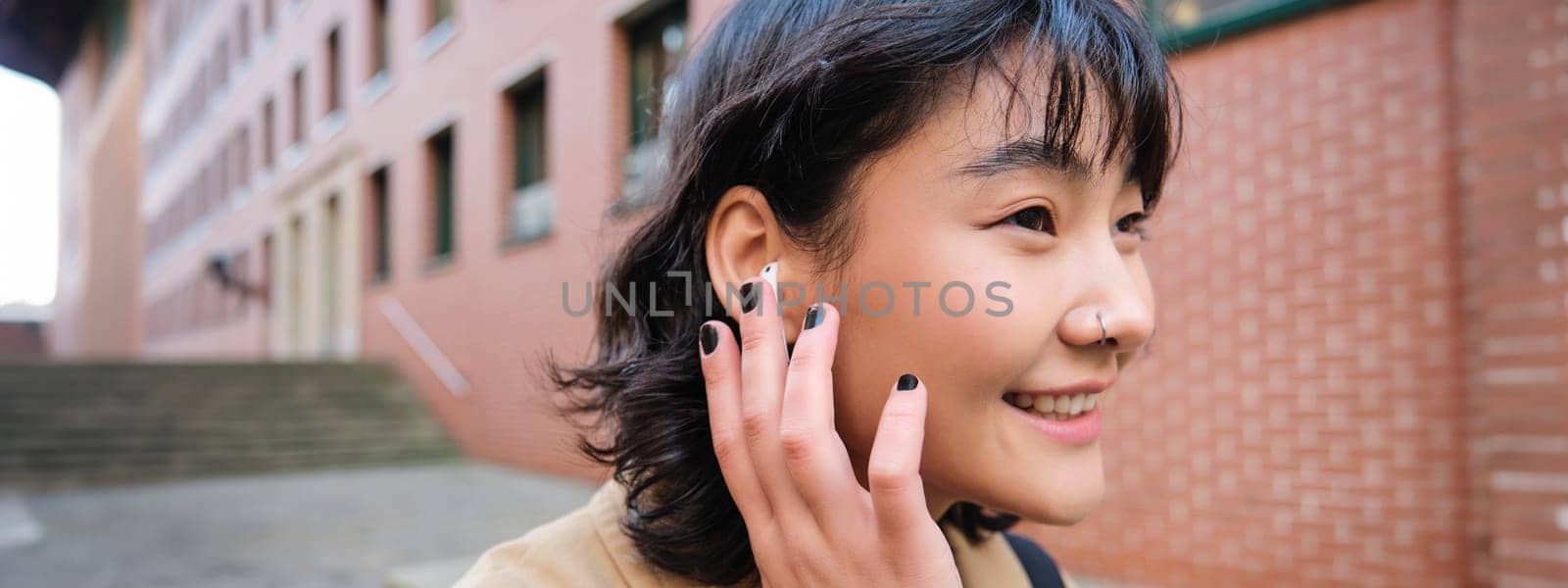 Close up portrait of young korean woman touches earphone, listens music in headphones with pleased smiling face, walks in city centre along the street by Benzoix