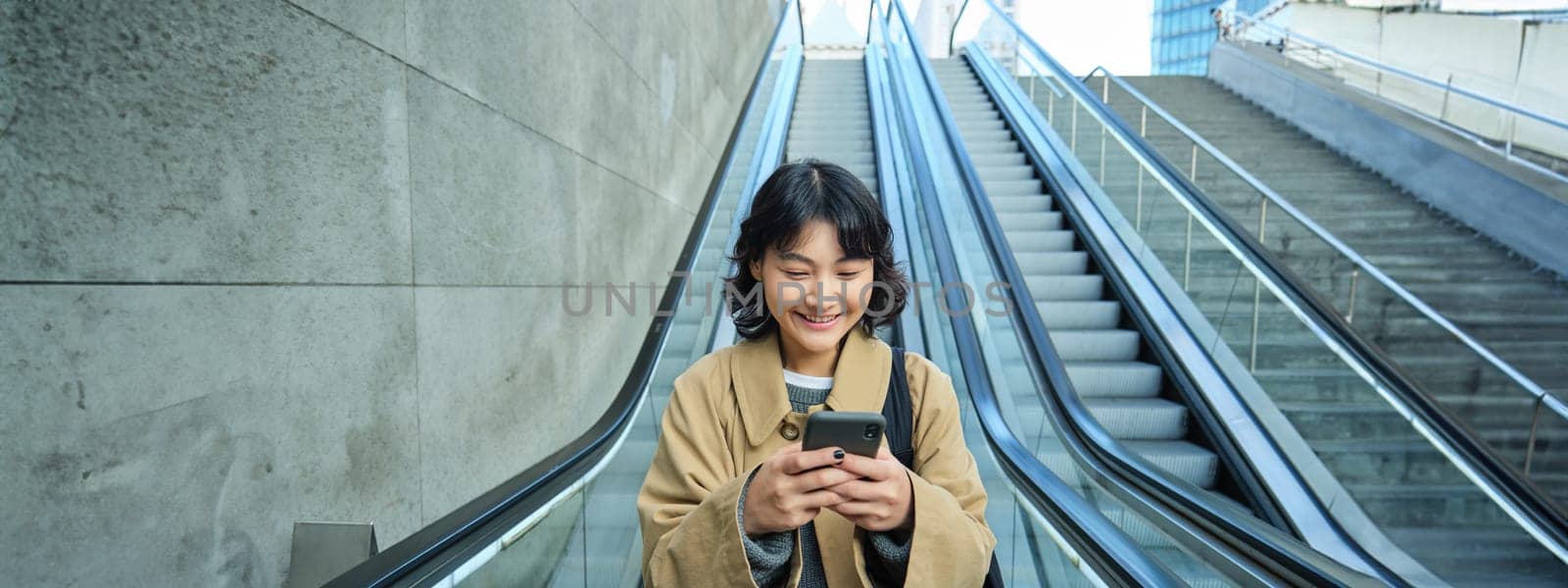 Portrait of beautiful asian girl, student goes down escalator in city, looks at mobile phone, uses telephone, map application, commutes somewhere in town by Benzoix