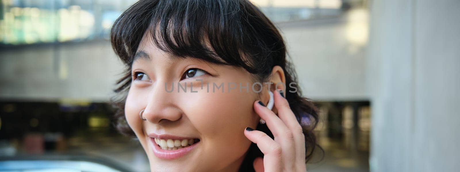 Close up portrait of smiling korean girl in headphones, listens music in wireless earphones, enjoys travelling around town with her favorite songs playlist by Benzoix