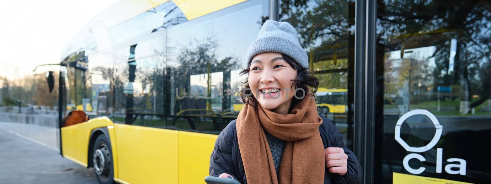 Portrait of girl standing near bus on a stop, waiting for her public transport, schecks schedule on smartphone application, holds mobile phone, wears warm clothes.