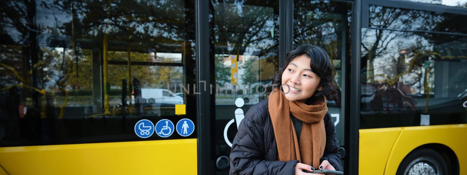 Portrait of korean girl looking for her bus on a stop, holding mobile phone, checking schedule, time table on smartphone app by Benzoix