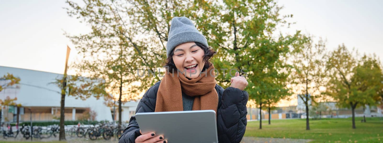 Portrait of happy asian girl sits on bench, looks at digital tablet screen and cheers, triumphs, wins something, celebrates good news, relaxes in park outdoors by Benzoix