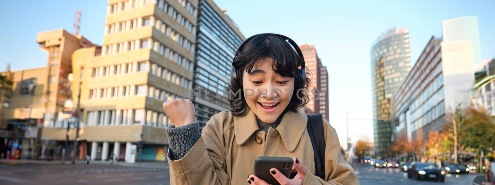 Young happy woman celebrating on street, holding smartphone and cheering, reacts amazed to good news, reads phone text message with surprised joyful face by Benzoix