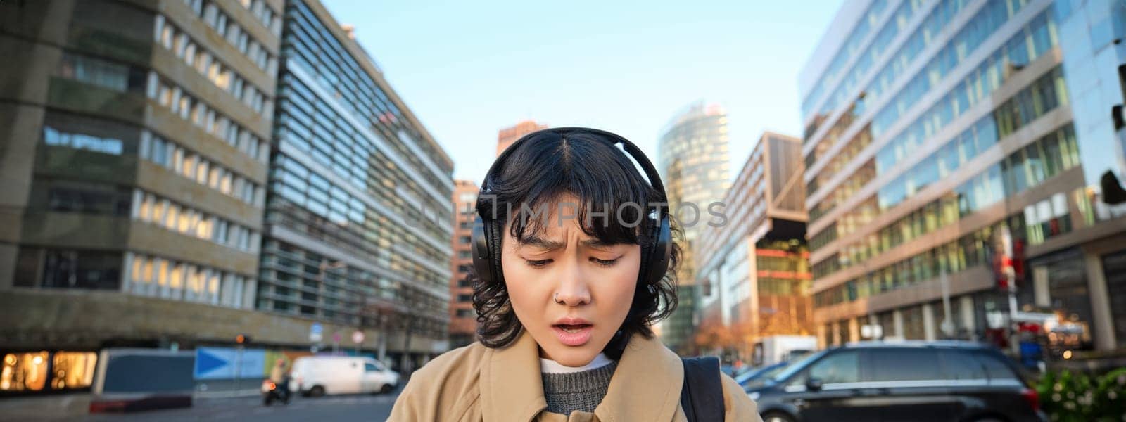 Portrait of young asian girl, student walks in city, listens music in headphones and uses mobile phone on streets, looks concerned at screen, reading bad news by Benzoix