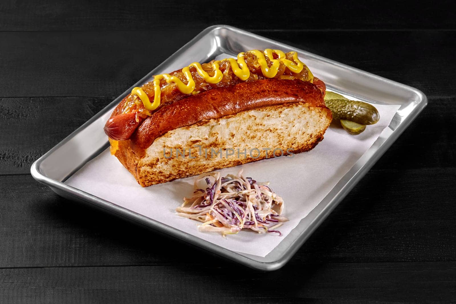 Delicious hot dog in toasted brioche sandwich bread with smoked sausage, caramelized onions and mustard sauce served on metal tray with pickled gherkins and shredded cabbage. Popular snack