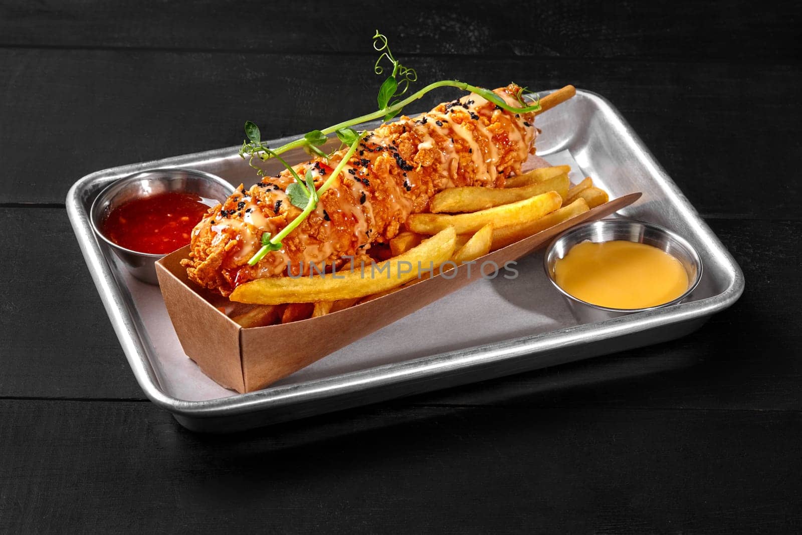 Delicious corndog with smoked sausage and crispy thick cornmeal batter on wooden skewer garnished with soil of grounded olives and microgreens served with potato fries and two sauces on metal tray