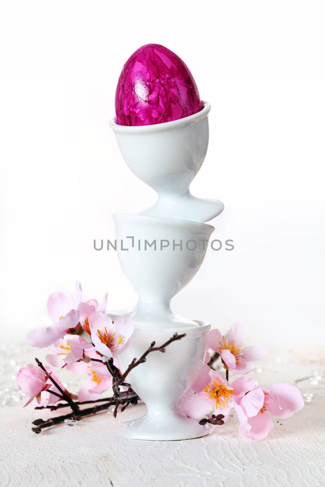 Three plain white china eggcups balanced precriously one on top of the other with a pink marbled Easter Egg and pink blossom.