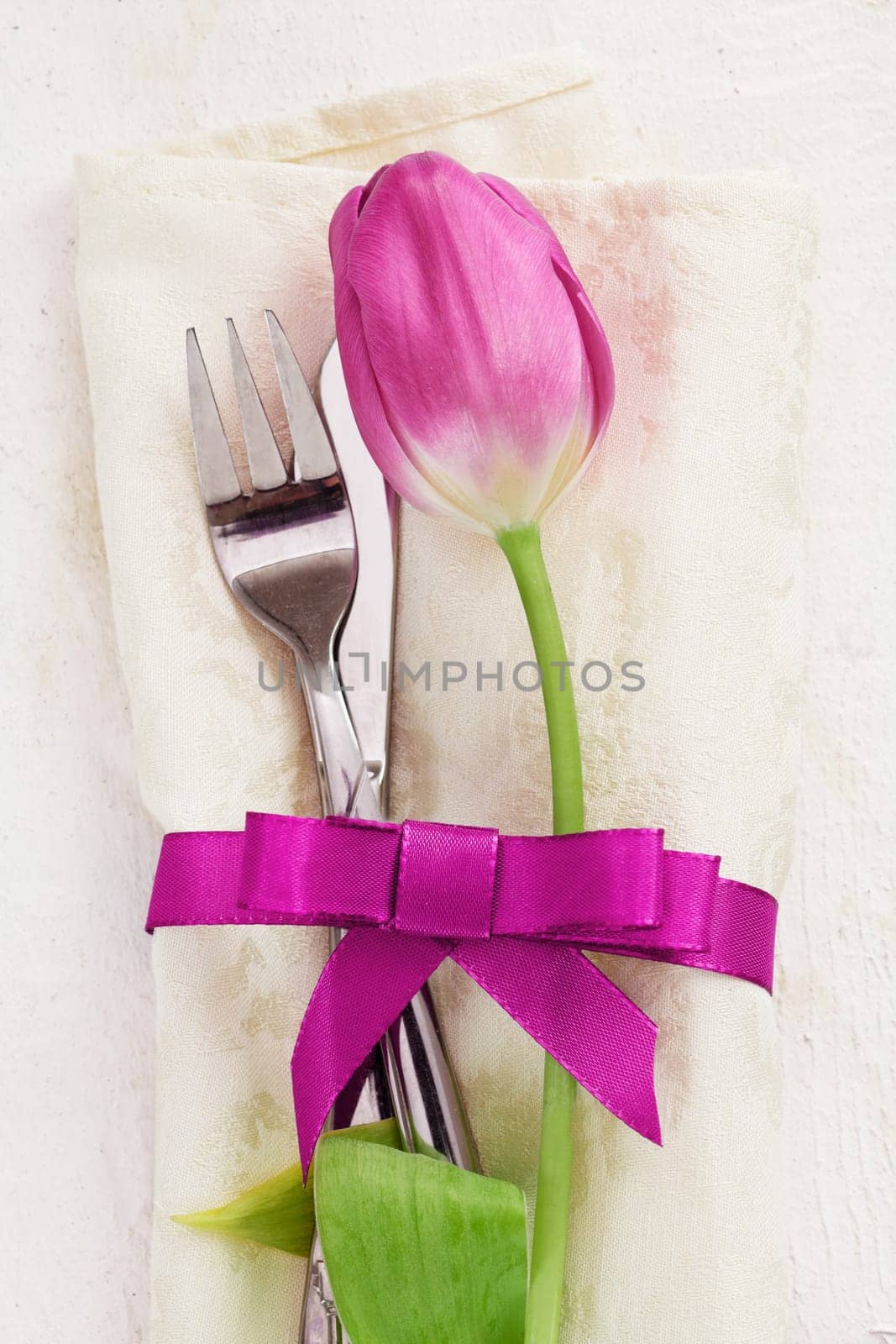 Napkin with tulip Flower. Dinner Table setting for two with a pink ribbon and a fork and a knife on wooden white background