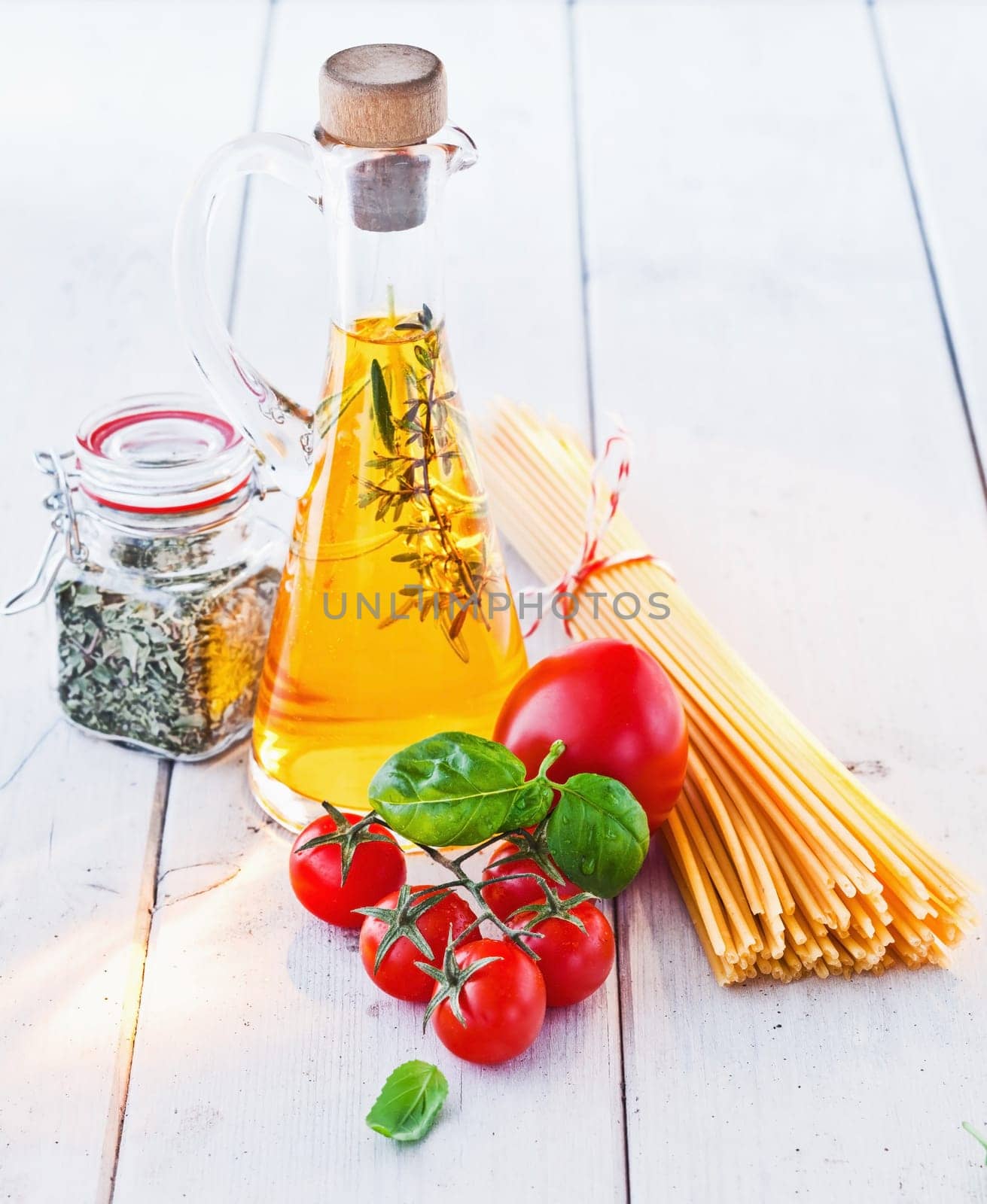 High angle view of fresh tomatoes and basil with uncooked spaghetti pasta and a flask of golden olive oil on a white painted wooden surface.