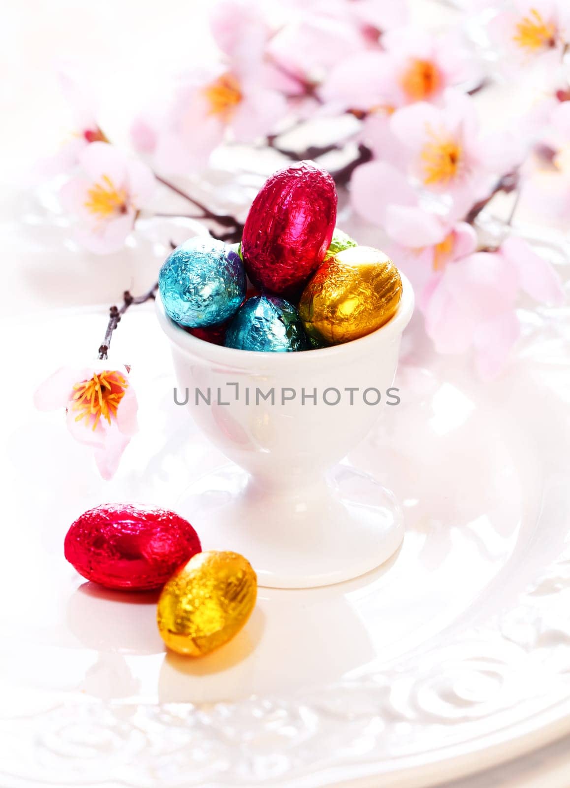 An eggcup full of tiny colourful foil wrapped chocolate Easter Eggs with cherry blossom