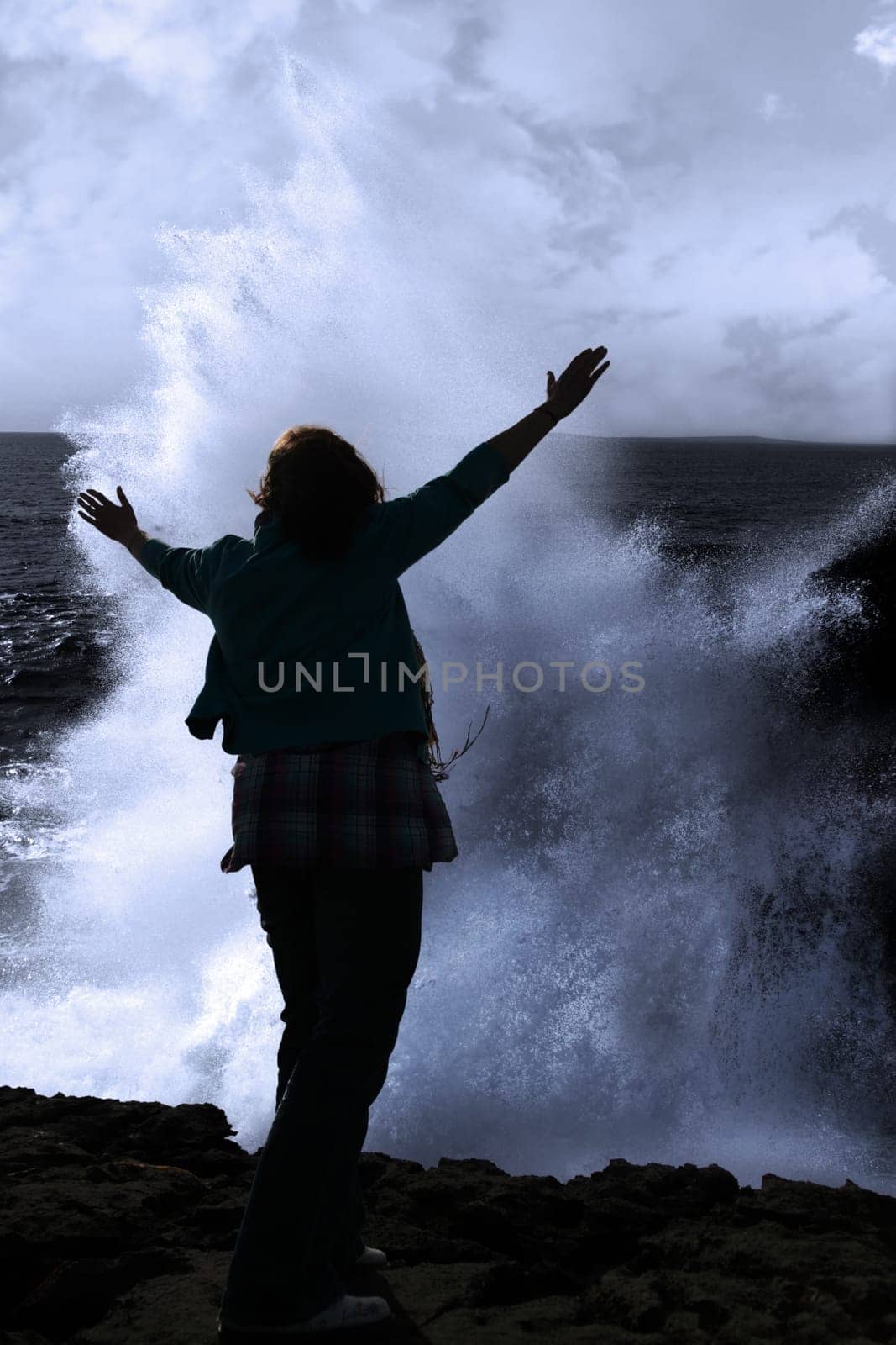 a lone woman raising her arms in awe at the powerful waves on the cliffs edge in county clare ireland