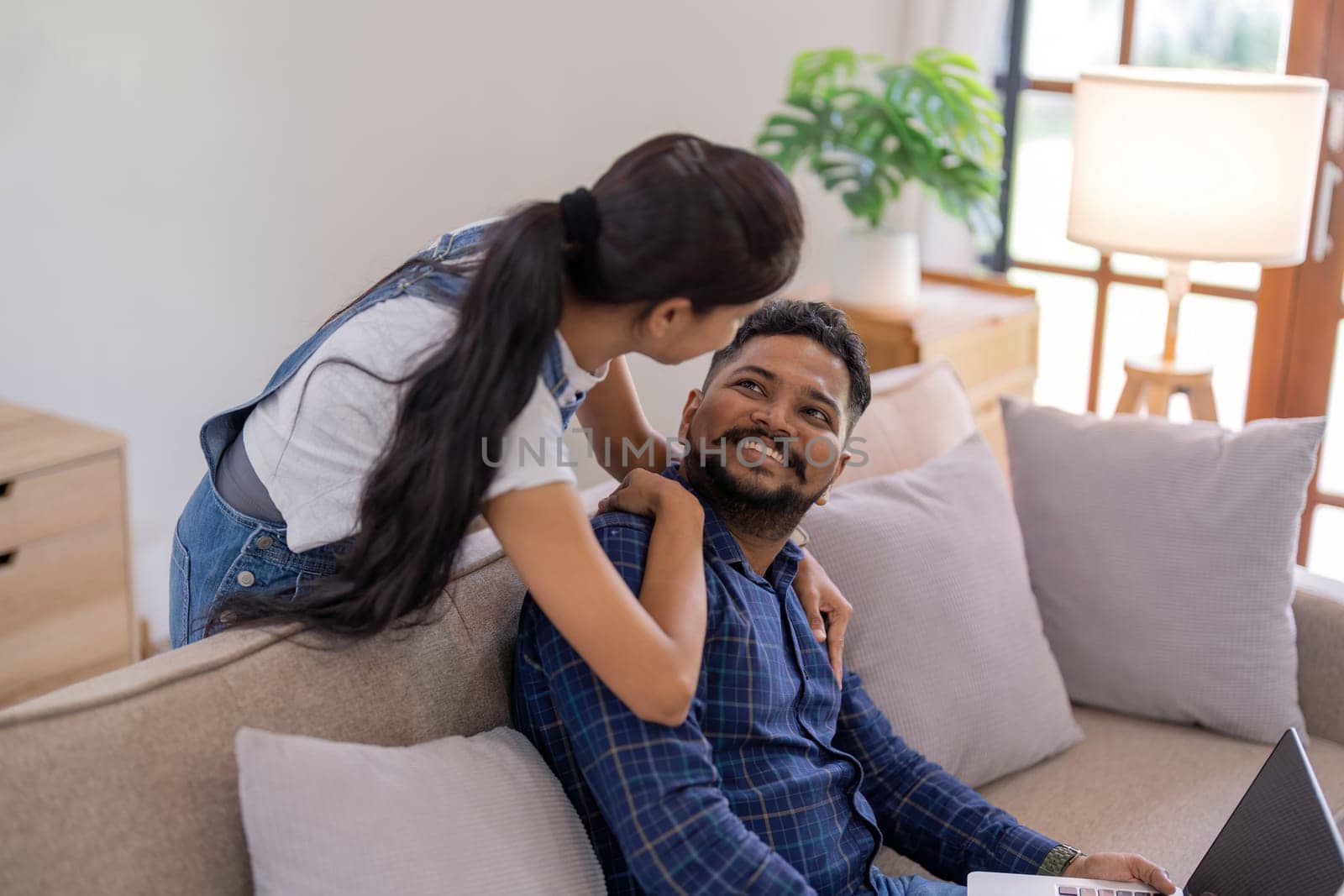 Happy lovely young Indian couple together at home, young wife hugging from behind her husband, sitting and rest on sofa in home, portrait of romantic multiracial couple in love by nateemee