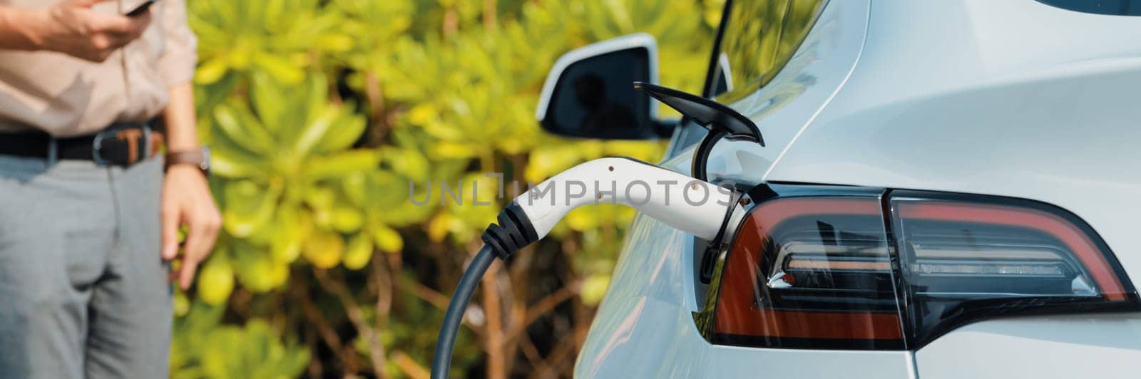 Young man use smartphone to pay for electricity at public EV car charging station green city park. Modern environmental and sustainable urban lifestyle with EV vehicle. Panorama Expedient
