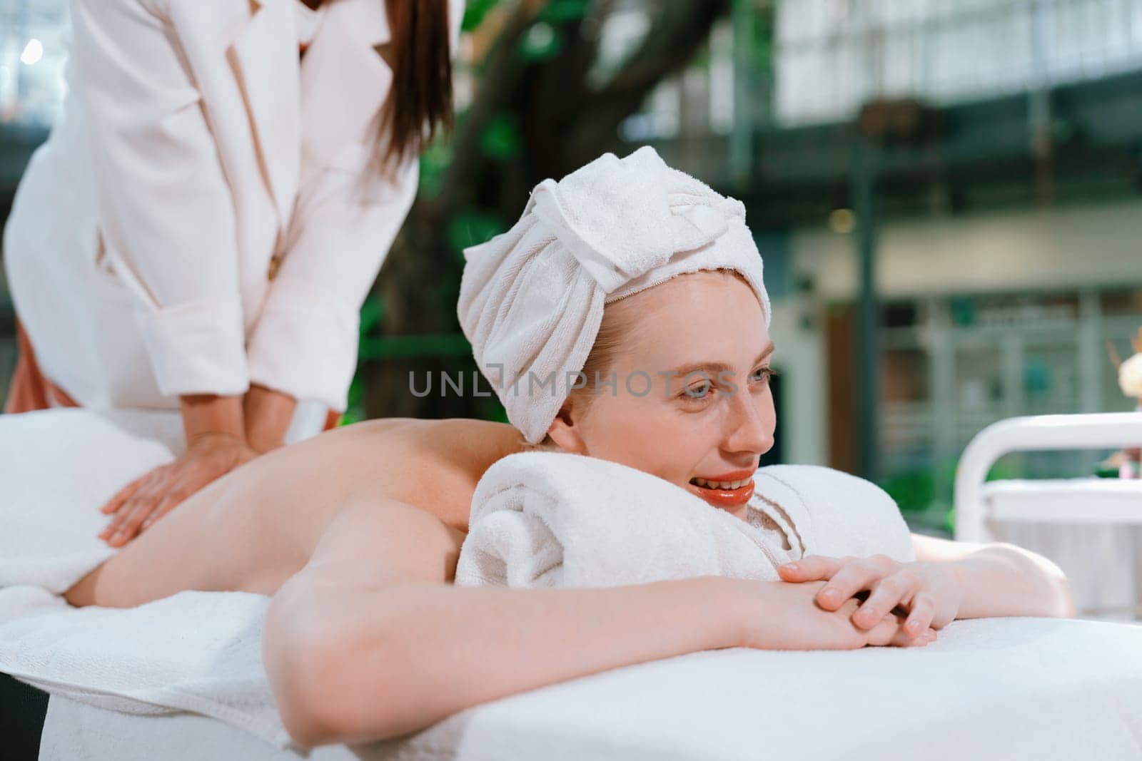 Beautiful young woman received a back massage on a spa bed from masseuse. Attractive female relaxes deeply by skilled hands of the massage therapist. Surrounded with nature. Side view. Tranquility.