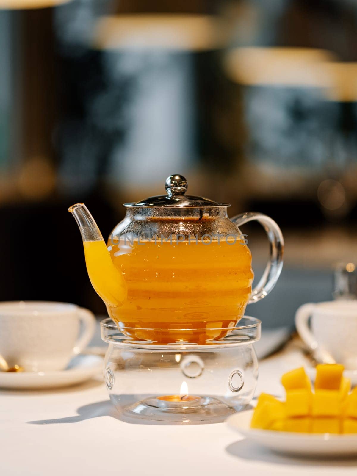 Mango herbal tea in teapot with warmer on table by fascinadora