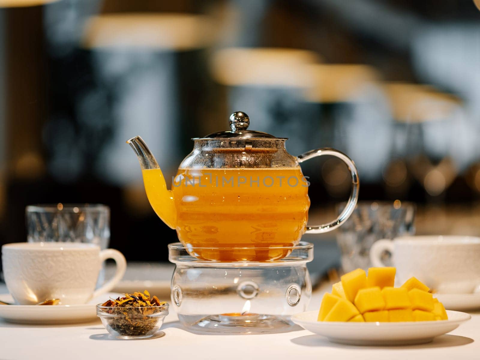 Herbal tea with mango fruit in transparent glass teapot with warmer and candle on table in restaurant. Herbal tea concept, wellbeing and health care