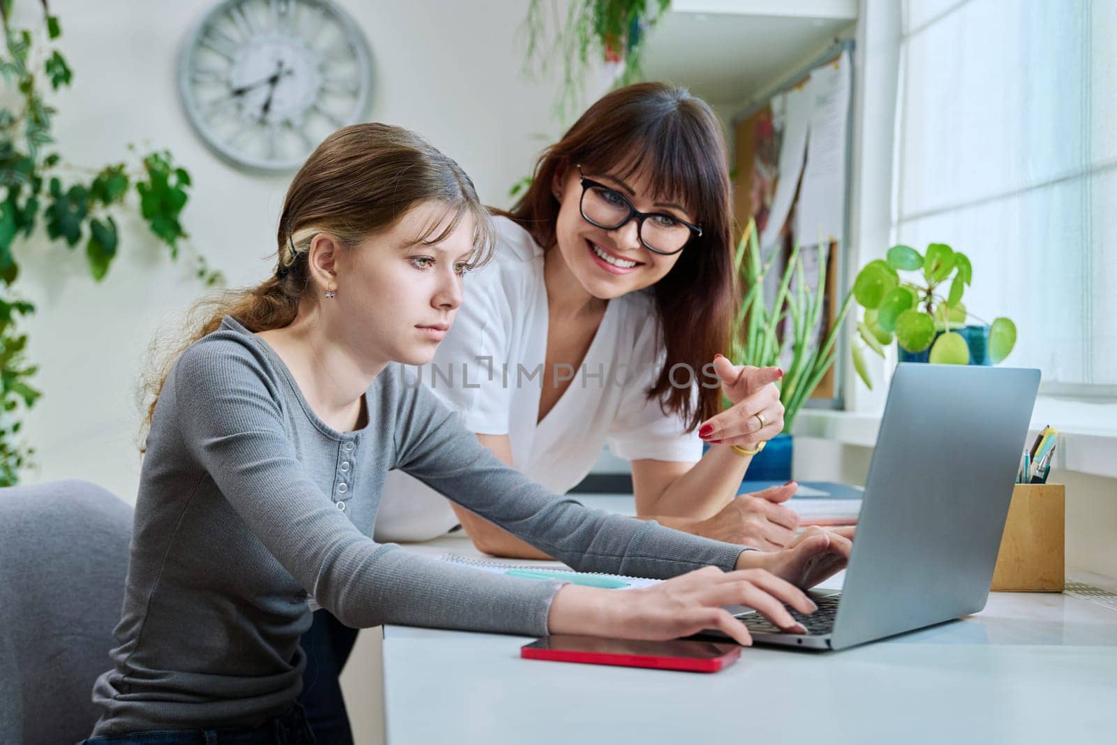 Mother and preteen daughter looking into laptop computer together, girl typing on keyboard, sitting at desk at home. Family, home lifestyle, study, kids concept