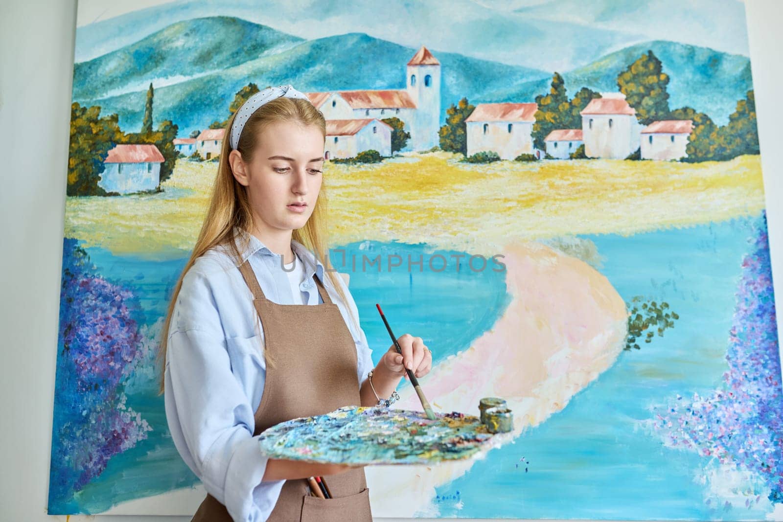 Young woman artist with acrylic paint palette with brushes painting picture on canvas on wall. Creativity, hobby, art therapy, leisure, paintings for sale, youth concept