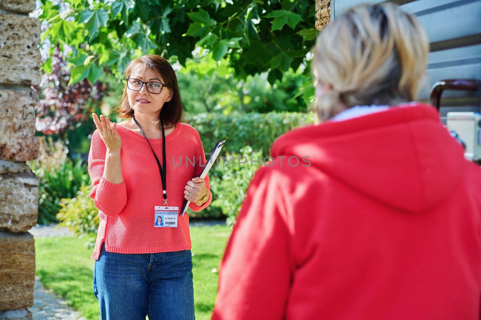 Female social counselor, health center worker, meets middle-aged woman near entrance gate to private backyard area