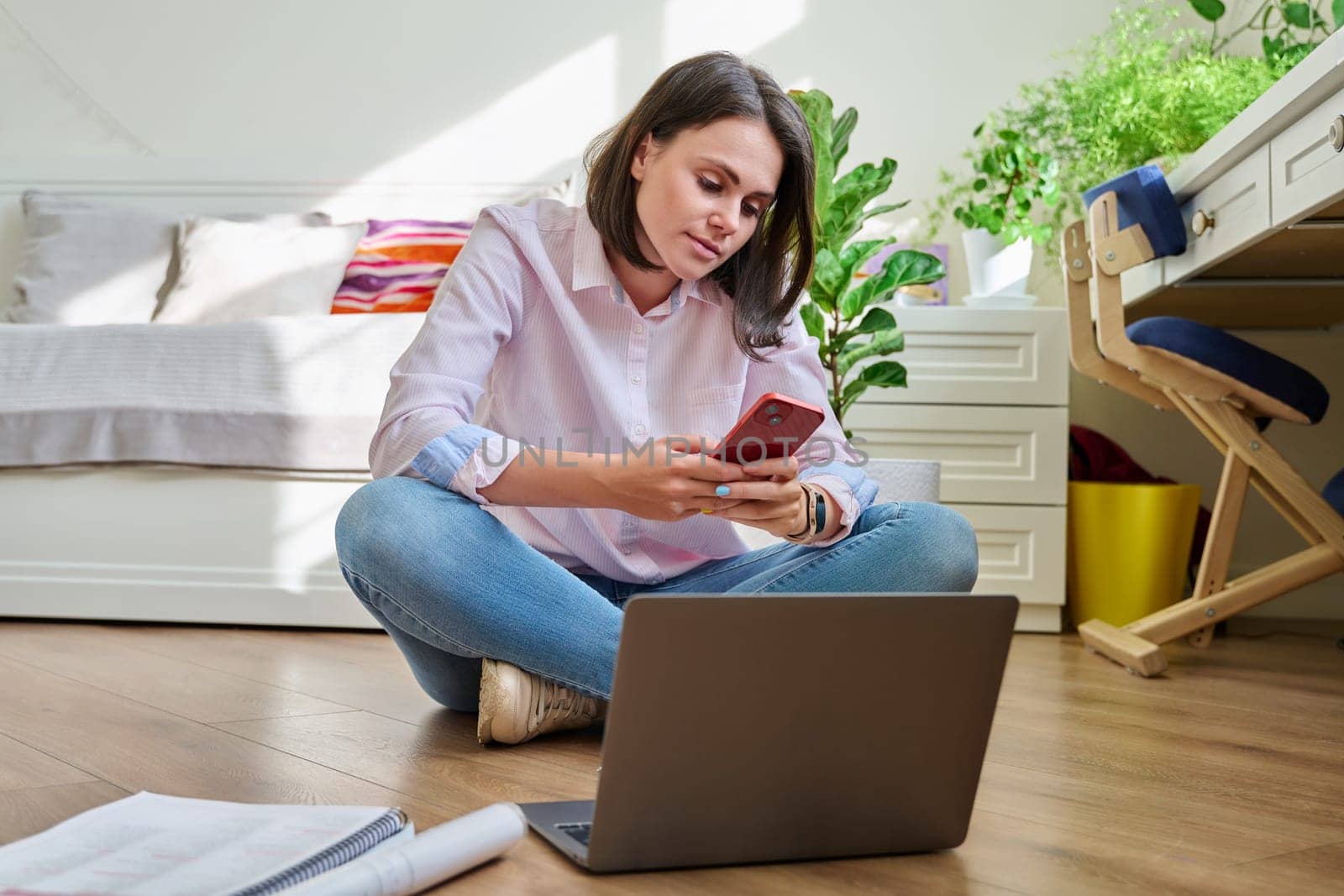 Young female university student studying at home sitting on floor using laptop phone by VH-studio