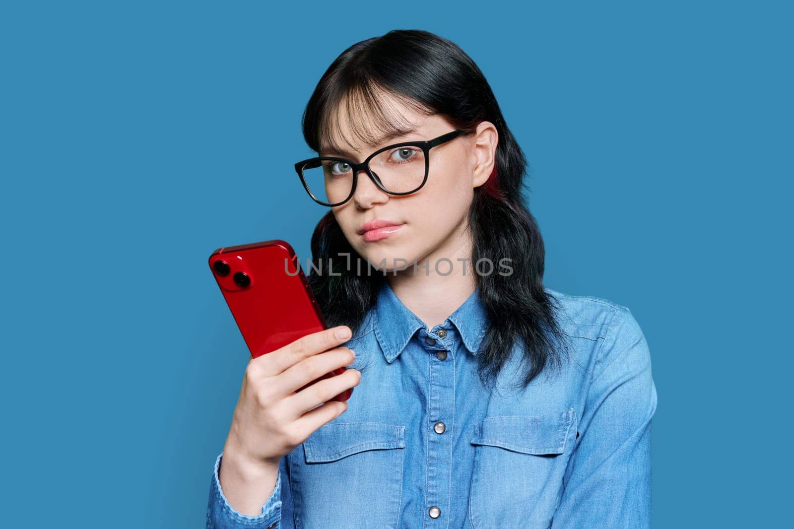 Serious teenage female with smartphone looking at camera on blue background by VH-studio