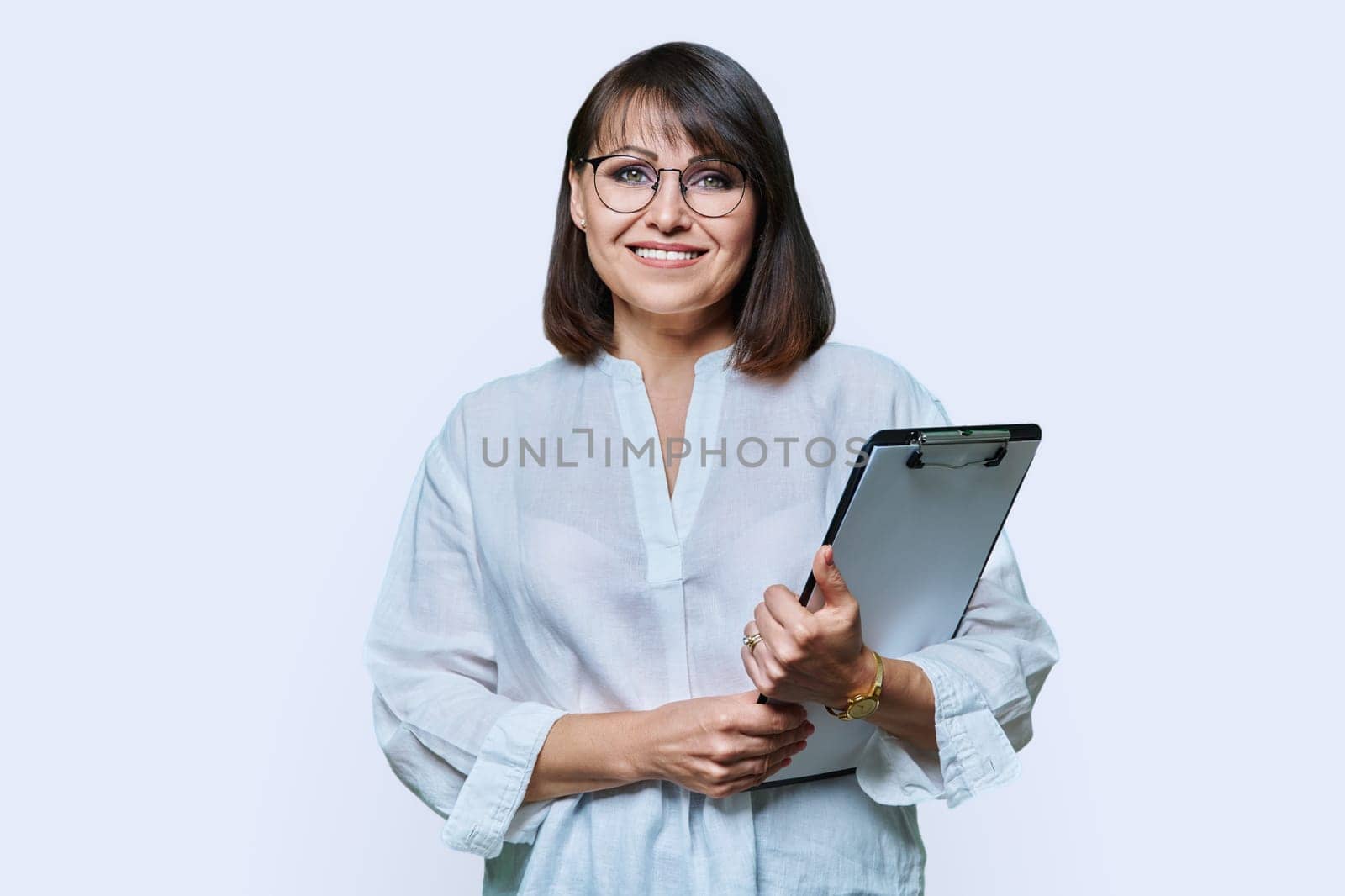 Confident middle aged business woman with clipboard looking at camera on white background. Mature smiling female teacher agent auditor psychologist supervisor posing with papers documents contracts