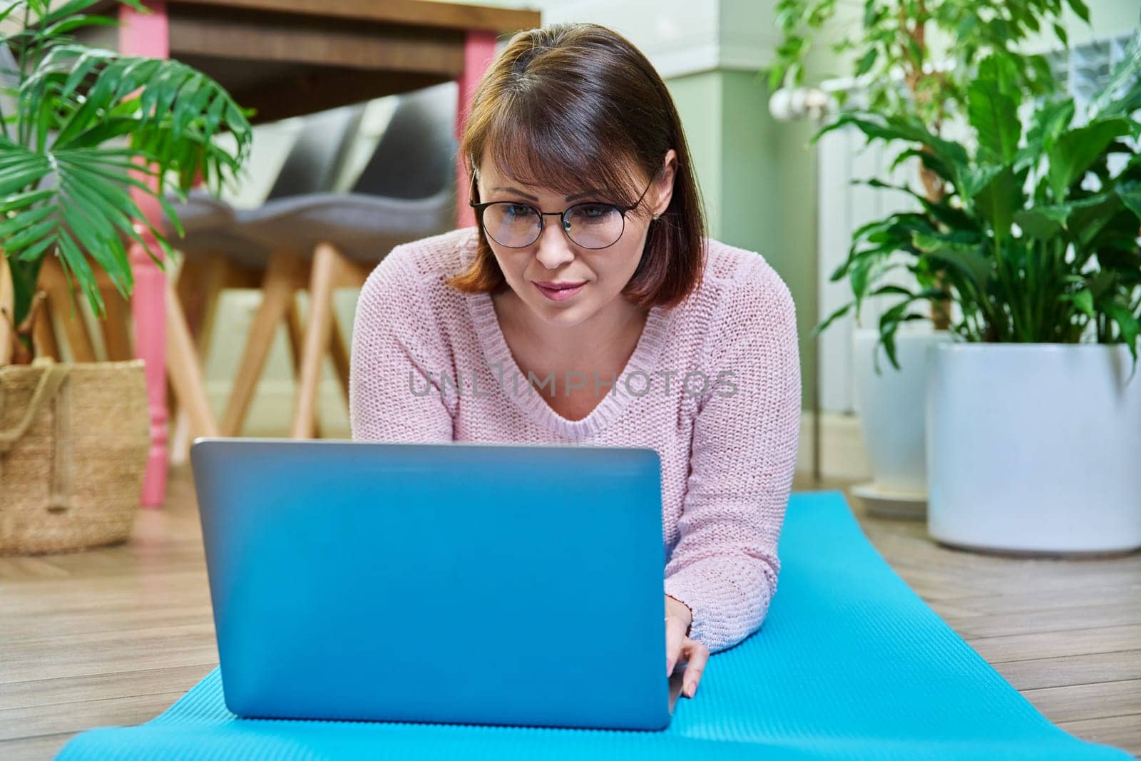 Middle-aged woman lying on a mat at home on the floor with a laptop. Lifestyle, leisure, people concept