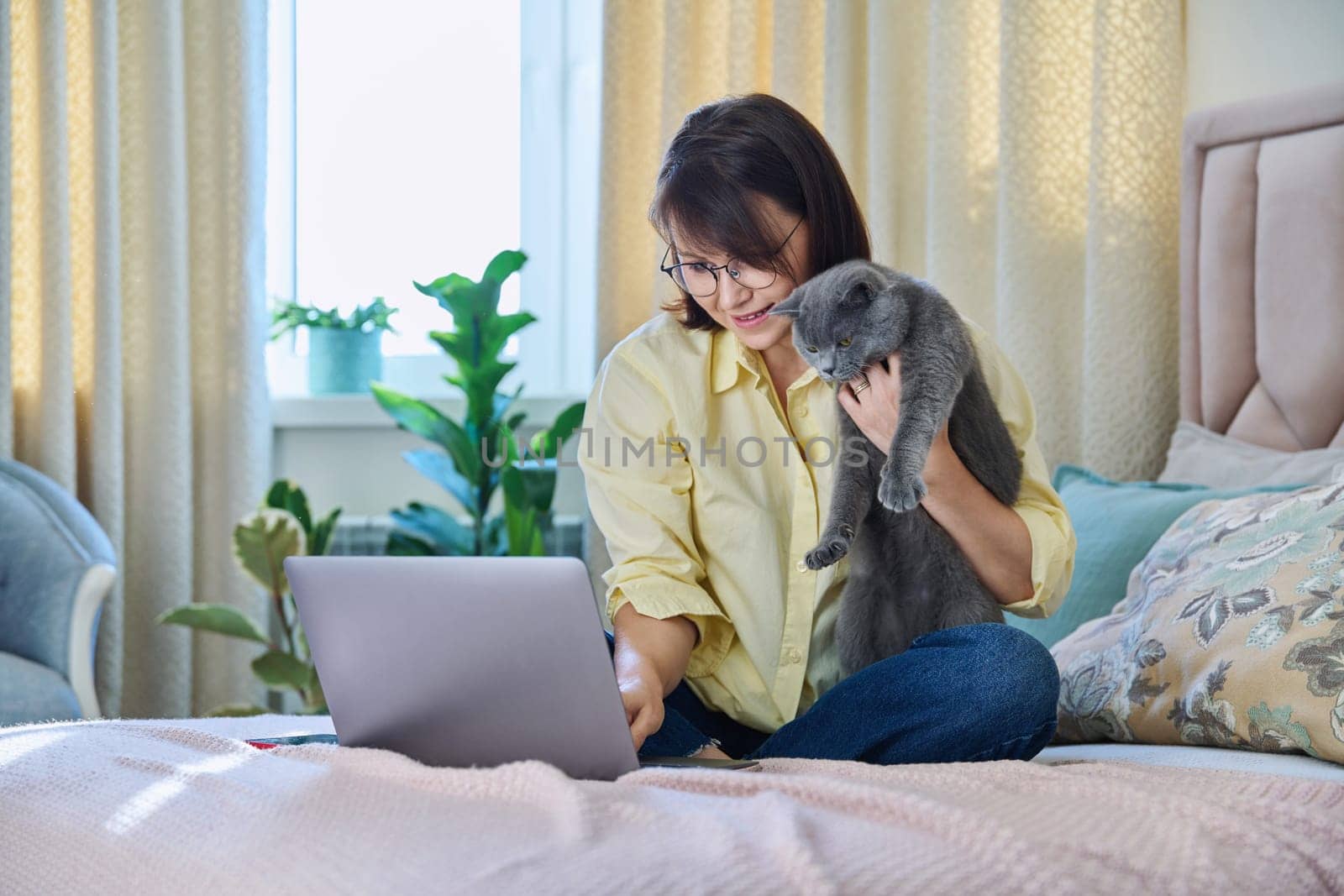 Middle aged woman at home on couch with laptop and cat by VH-studio