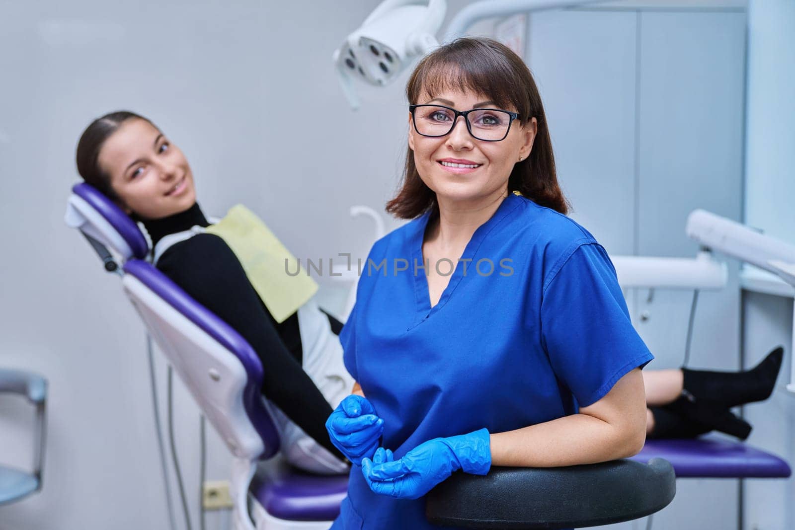 Portrait of smiling female dentist looking at camera with young teenage girl patient sitting in dental chair. Visit to dentist examination treatment. Dentistry hygiene dental teeth health care concept