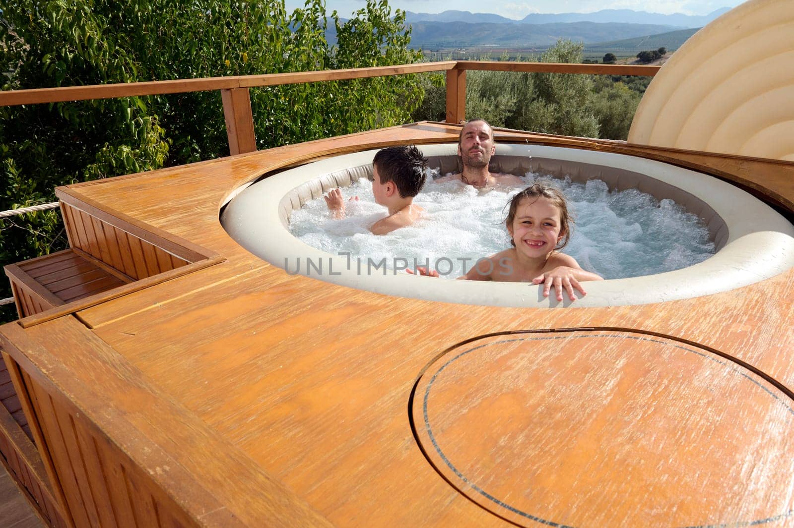 Cheerful mischievous little girl playing in the swimming pool with her brother and dad, smiles looking at camera by artgf