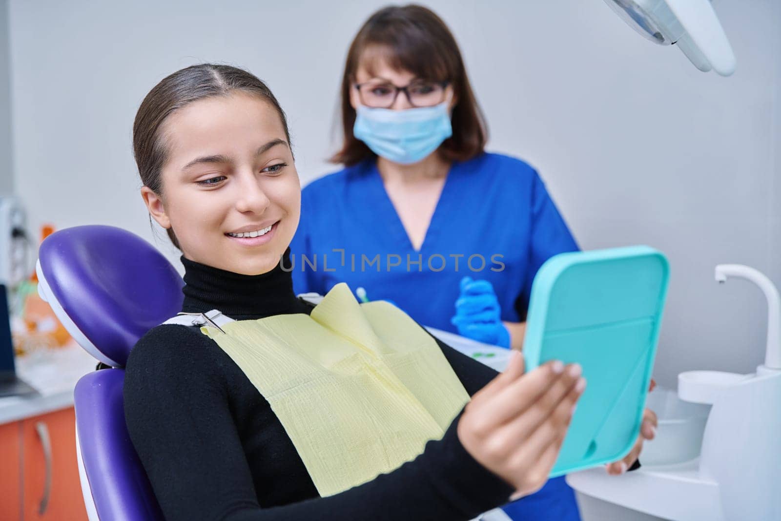 Happy young teenage female with mirror sitting in dentist chair smiling looking at healthy teeth, with dentist doctor in office. Dentistry, hygiene, treatment, dental health care concept