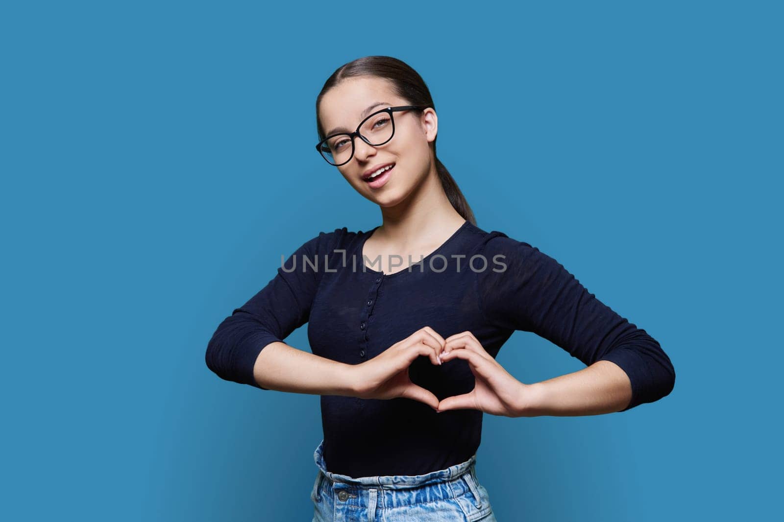 Young smiling female showing heart gesture with fingers, blue background by VH-studio