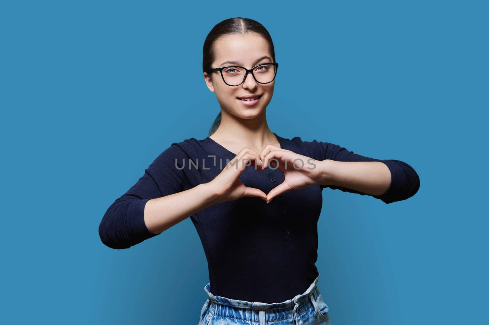 Young smiling female showing heart gesture with fingers, blue background by VH-studio