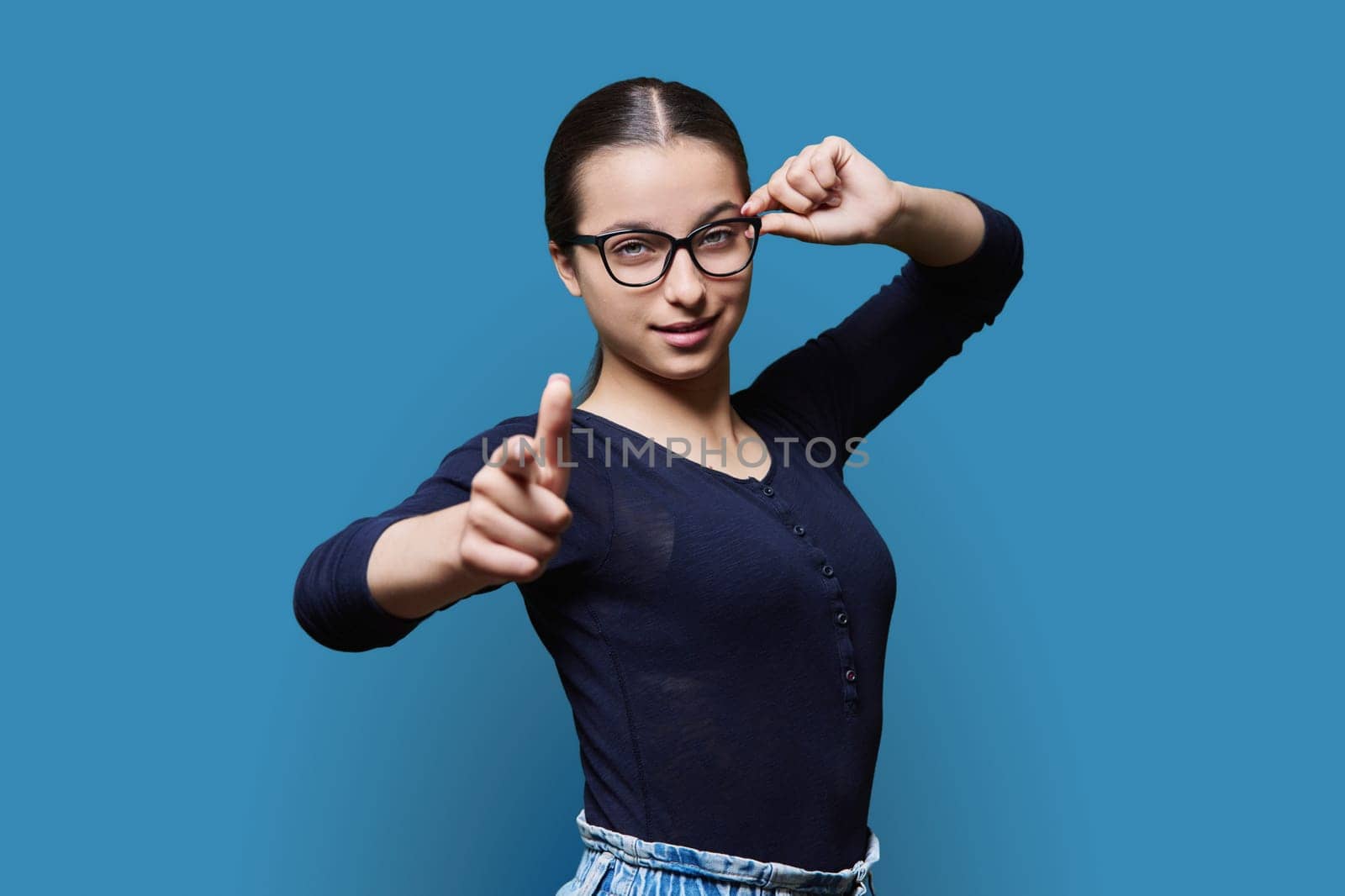 Teenage student girl in glasses looking at camera pointing finger at you, on blue color studio background. Announcement, highlighting, attention, education, lifestyle people concept