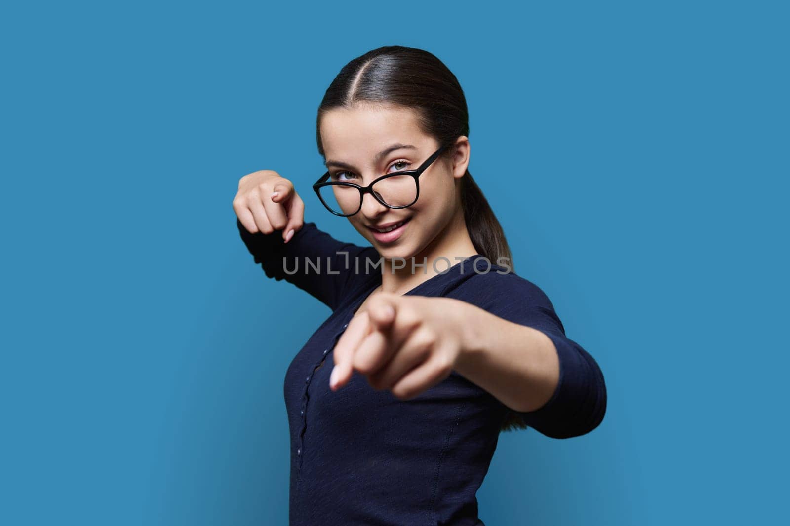 Teenage student girl in glasses looking at camera pointing finger at you, on blue color studio background. Announcement, highlighting, attention, education, lifestyle people concept