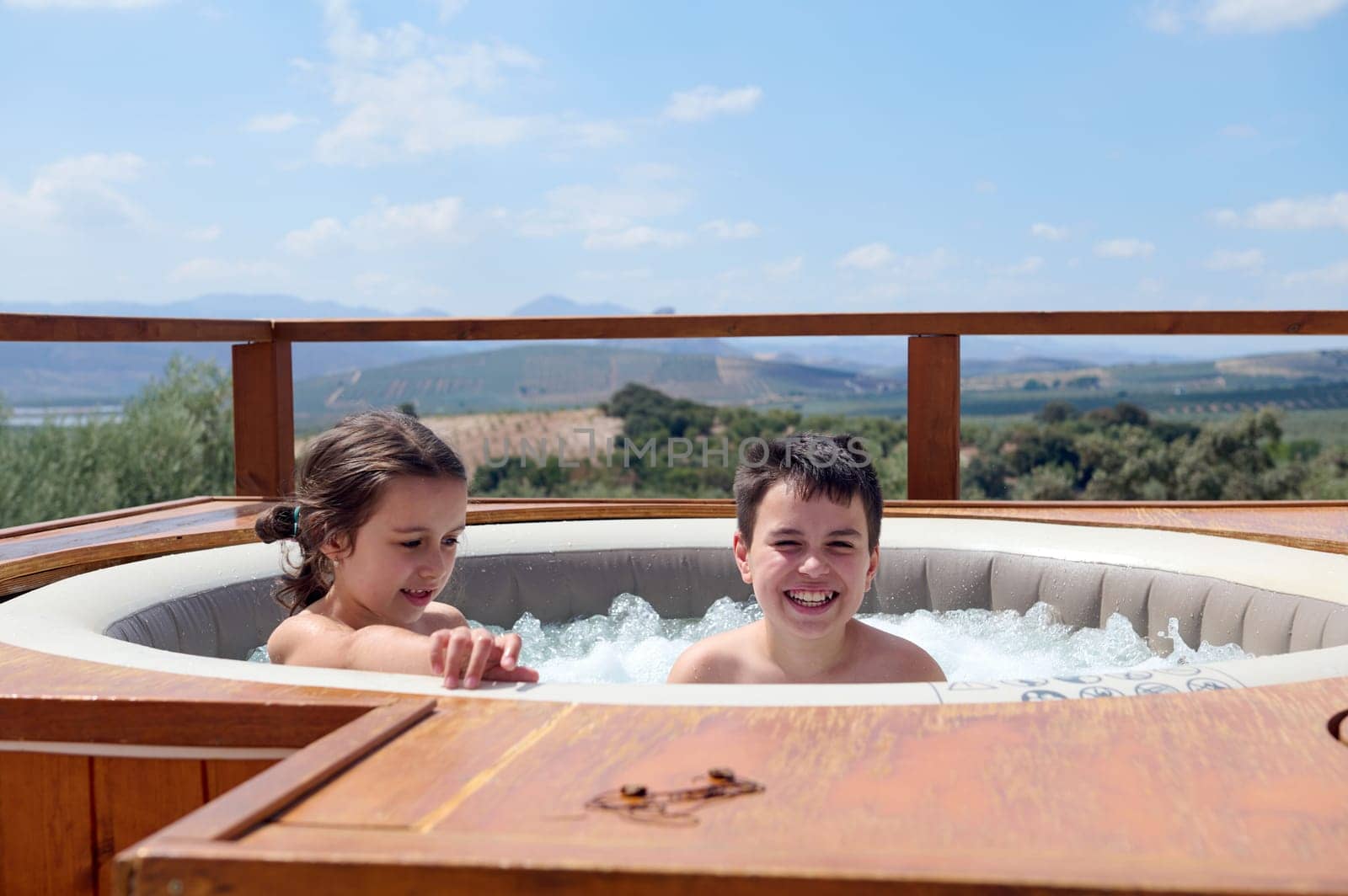 Happy Caucasian kids, a teenage boy and his little sister smiling looking at camera while playing in the swimming pool with bubbles, enjoying happy family holidays in Spain. People. Vacations