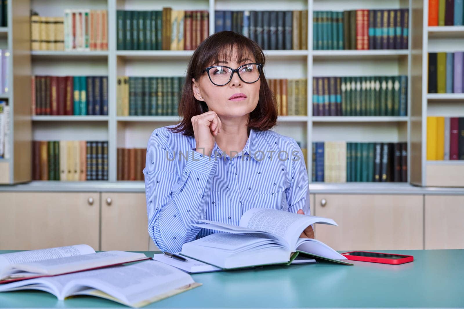 Middle-aged female teacher working in library, writing in papers at desk. Education, knowledge, school, college, university, technology, teaching concept