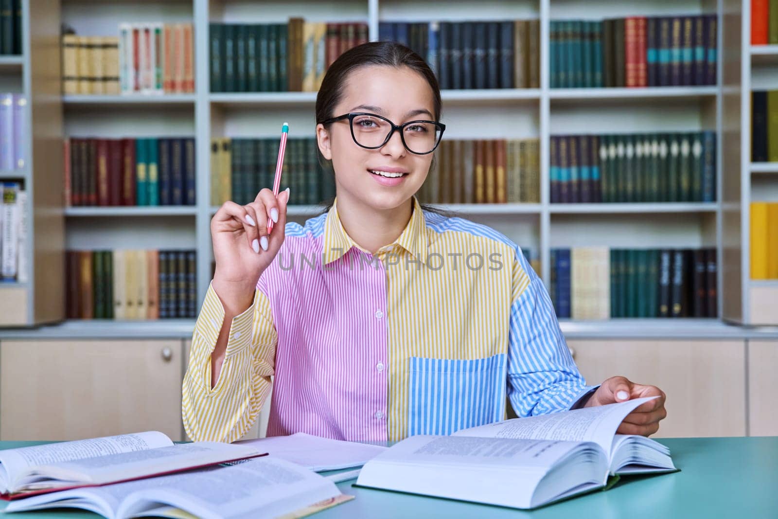 Teenage girl student studying in library. Female looking at camera, making notes in notebook. Adolescence, knowledge, education, high school concept