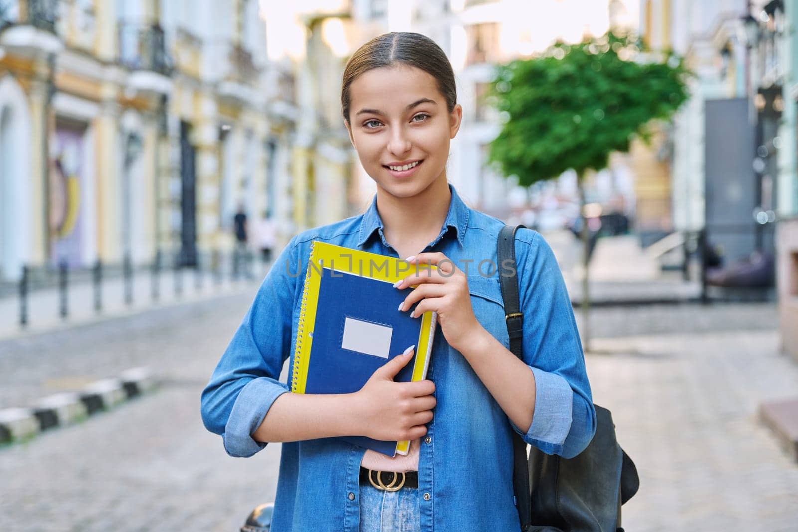 Outdoor portrait of smiling teenage female student looking at camera in city. Girl with backpack with books and notebooks in hands. Education, knowledge, high school, adolescence concept