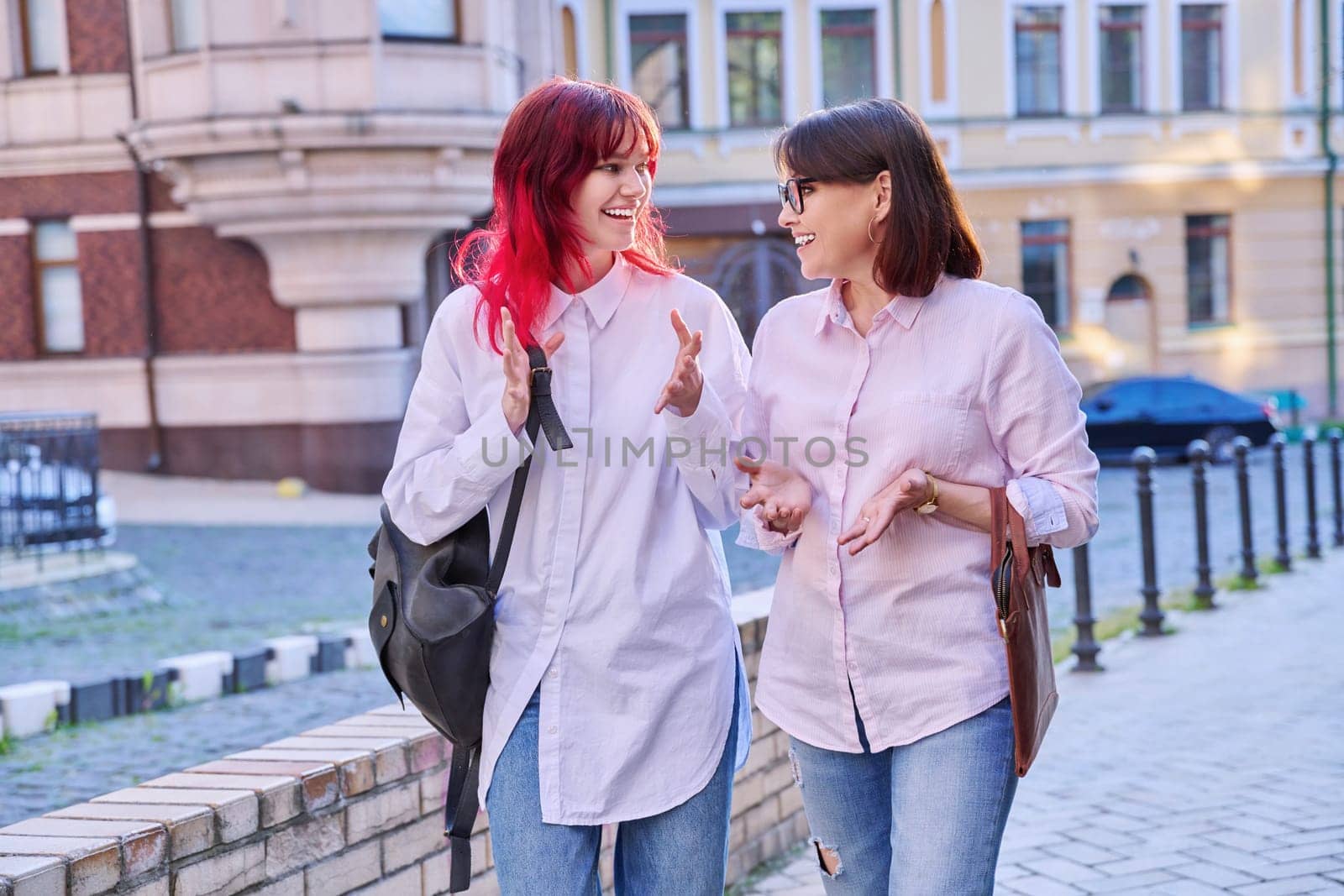 Two generations, smiling joyful happy mother and stylish attractive teenage daughter, walking talking together along city street. Family communication parent child teenager lifestyle leisure people