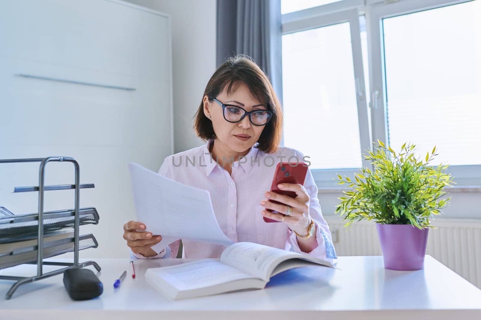 Middle aged business woman working at home sitting at the table with smartphone documents books. Freelance, remote work, home office concept