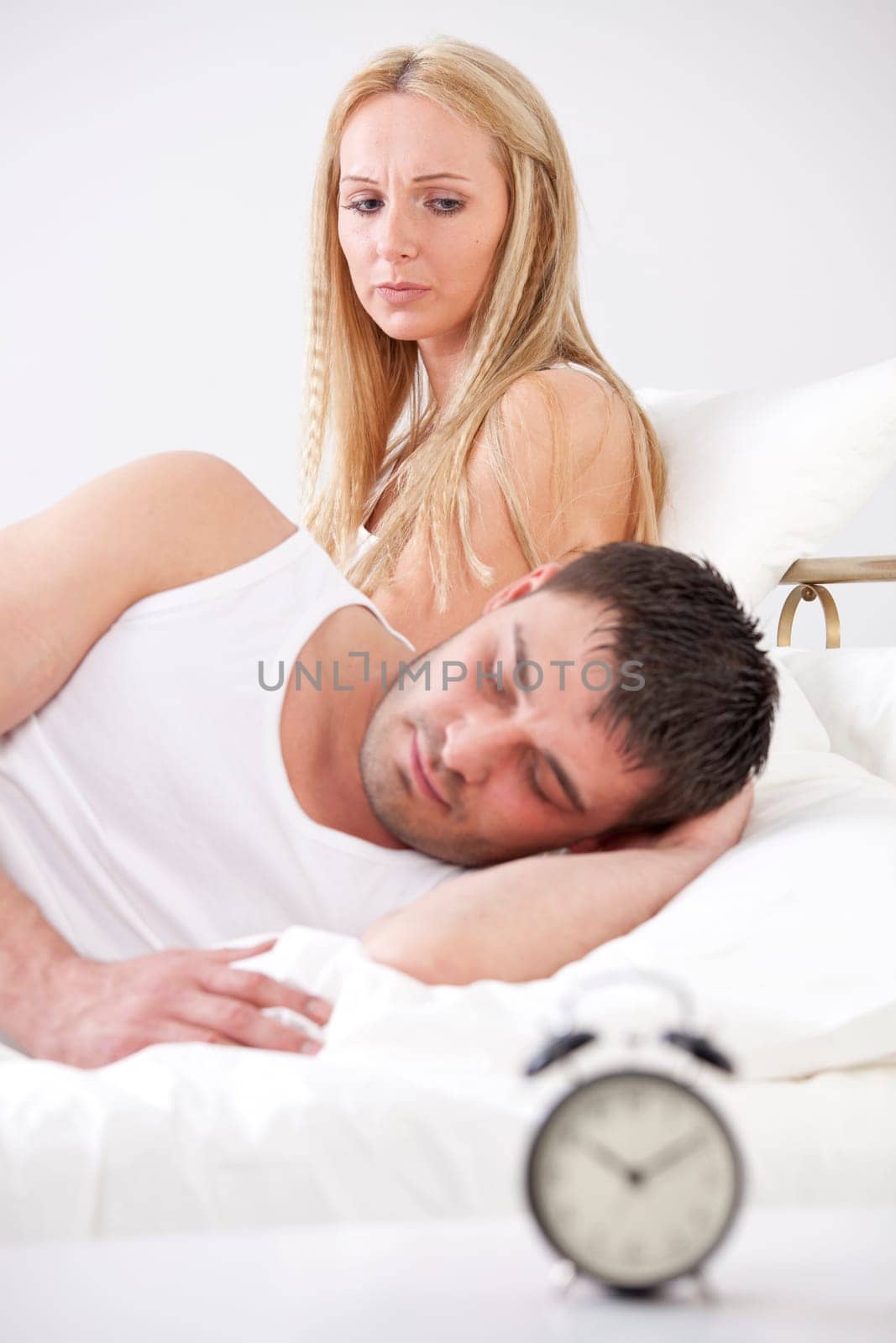 Frustrated woman in bed looking at sleeping man - alarm clock in foreground